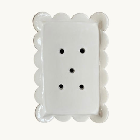 A white ceramic salt and pepper shaker plate designed like a biscotti cookie, featuring a scalloped edge and five perforations on the top surface, set against a solid off-white background.