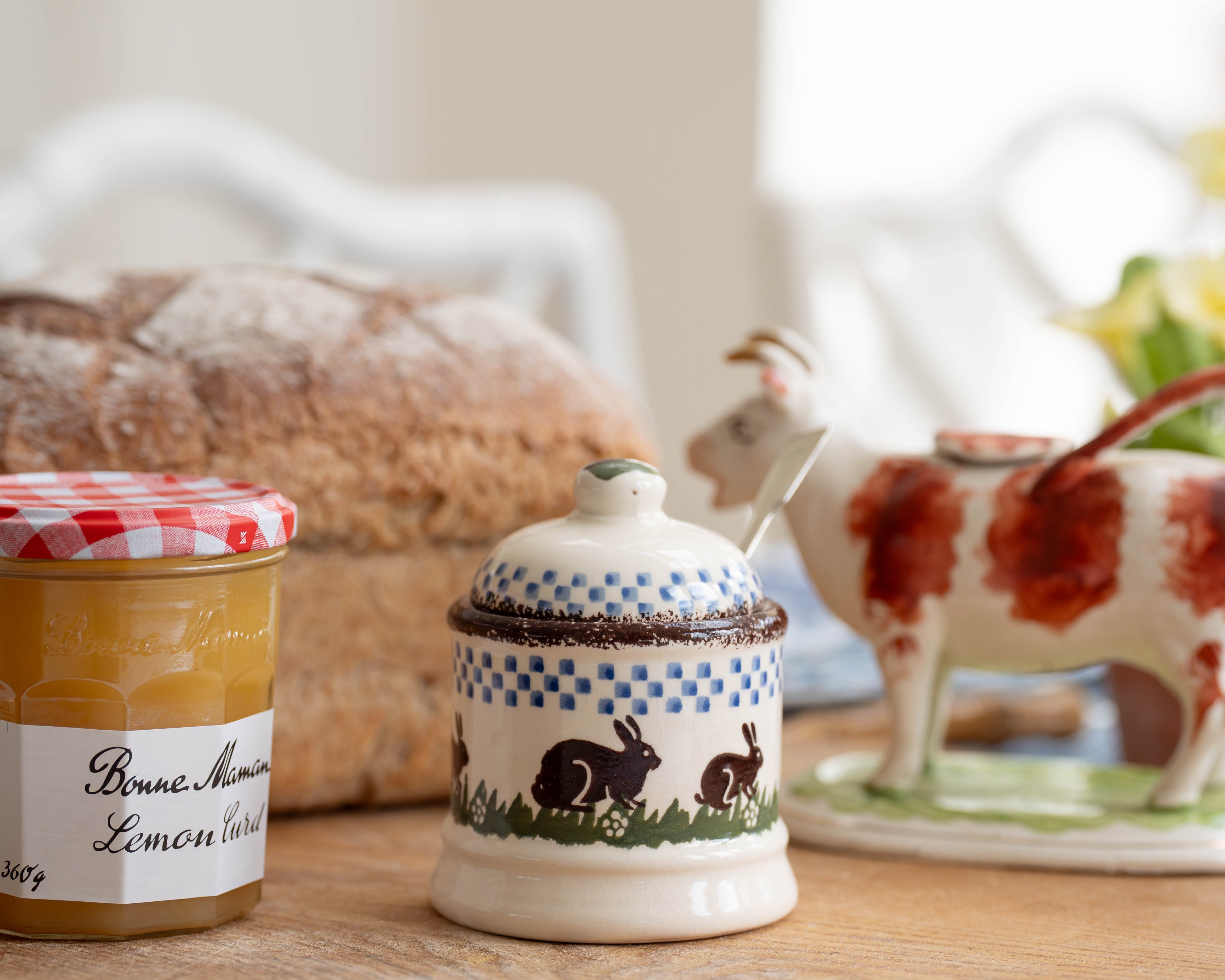 Two ceramic jars with lids on a wooden surface, with bread and a cow-shaped teapot in the background.